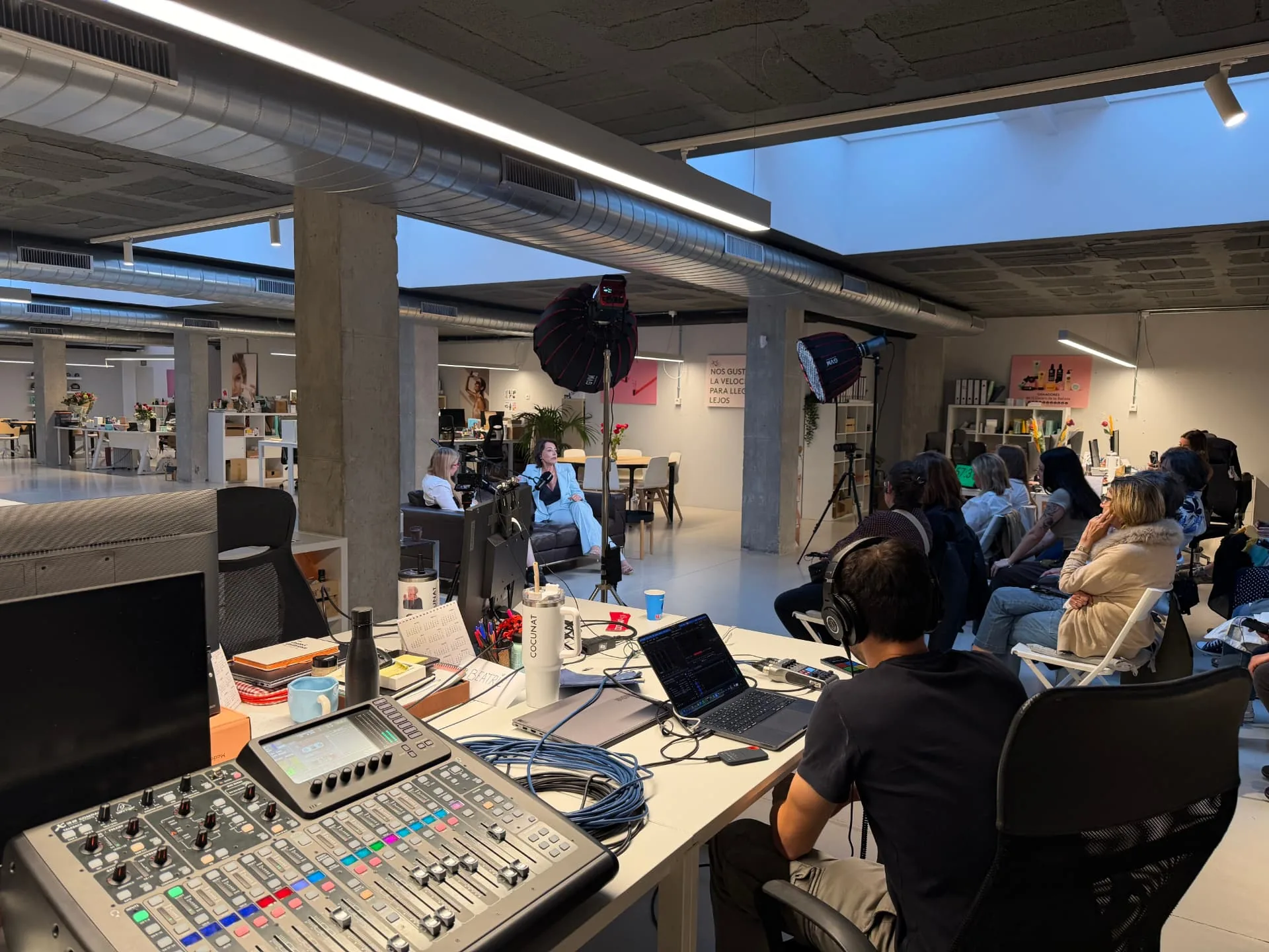 Grabación de un podcast en curso: mesa de control con técnico de sonido, dos personas entrevistándose en sillones frente a los focos y público asistente